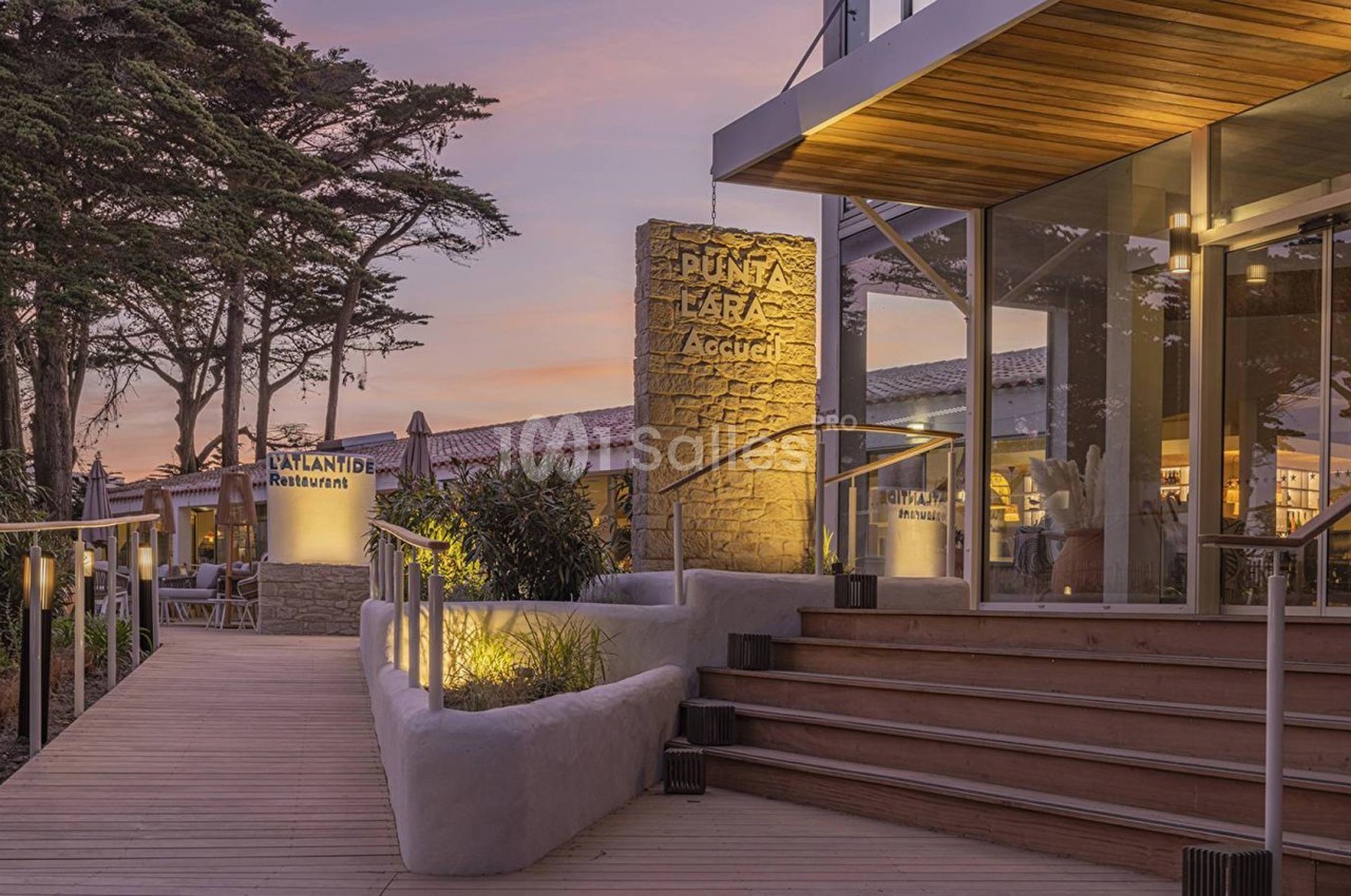 Entrée d'un bâtiment moderne avec terrasse en bois, éclairage nocturne et végétation, sous un ciel au coucher du soleil.