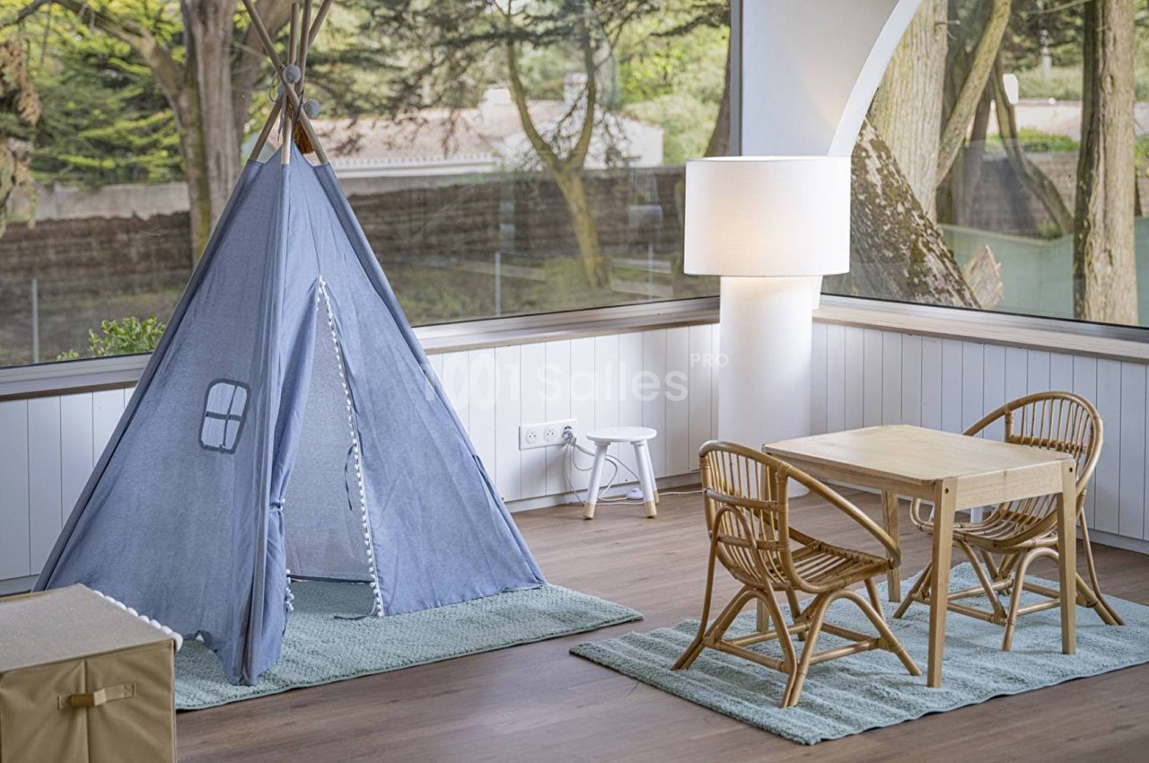 Tente bleue pour enfants et mobilier en rotin dans une pièce lumineuse avec vue sur un jardin boisé.