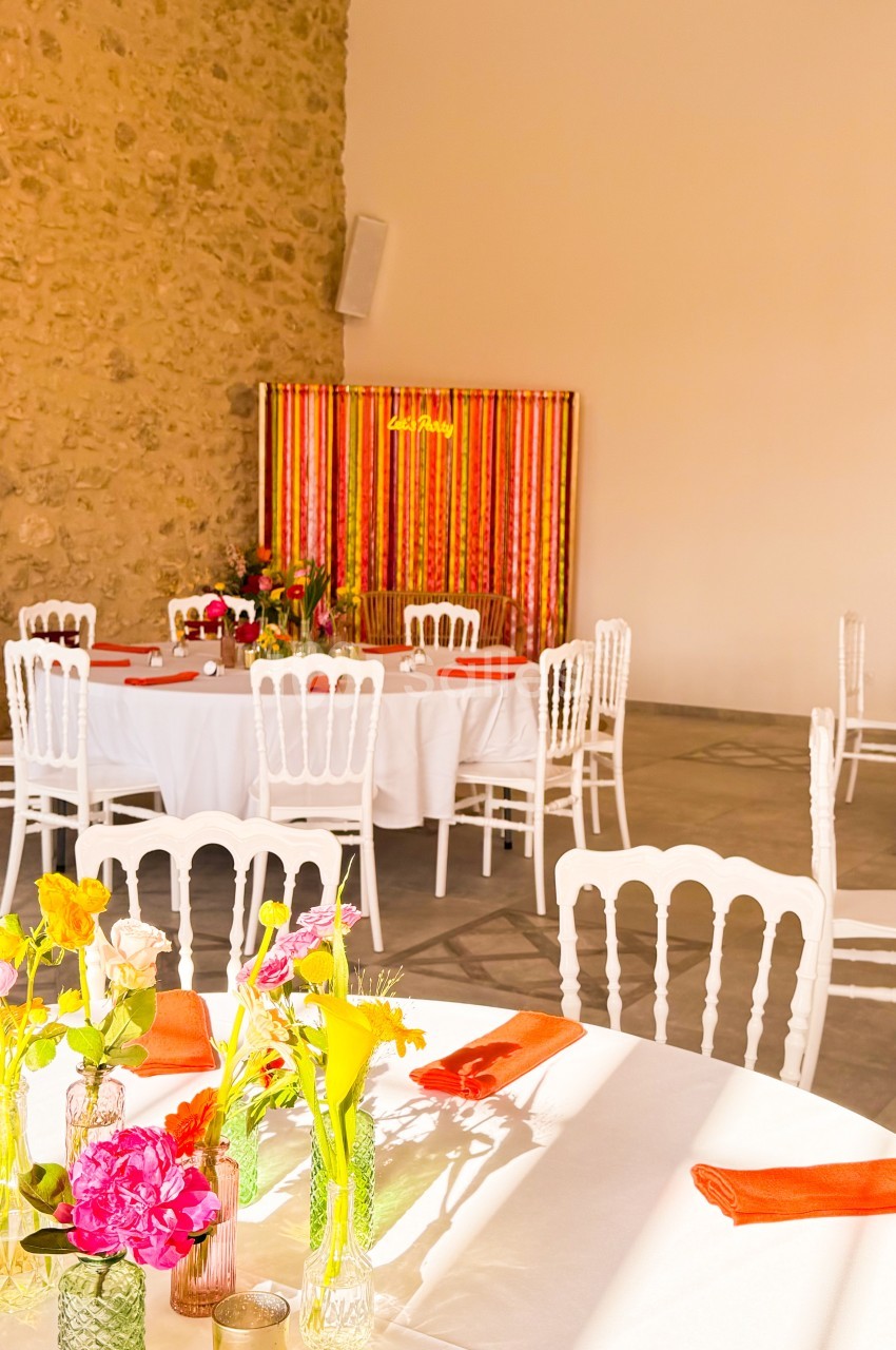 Salle décorée pour un événement avec tables rondes, nappes blanches, chaises blanches et fleurs colorées en centre de table.
