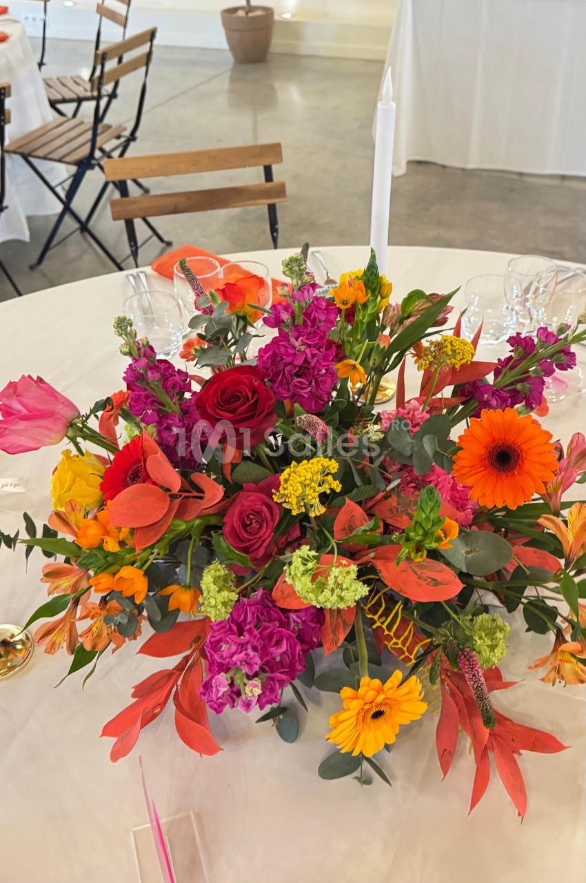 Centre de table floral coloré avec des roses rouges, des gerberas orange et divers feuillages sur une nappe blanche.