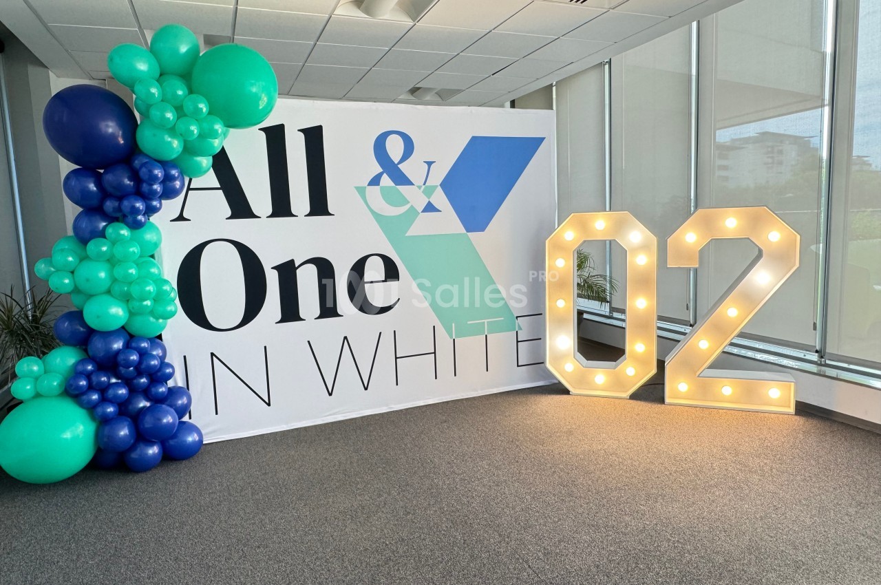 Mur décoratif avec ballons verts et bleus, panneau ’All & One in White’ et chiffre lumineux ’02’ dans un espace intérieur.