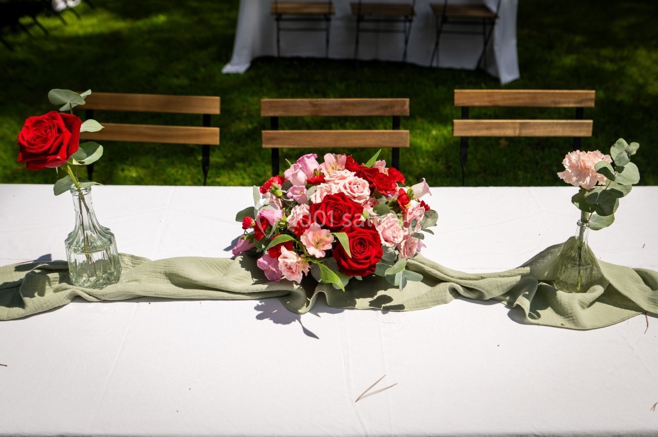 Bouquet de roses rouges et roses sur une table blanche décorée, entouré de vases avec des fleurs et un chemin de table vert.