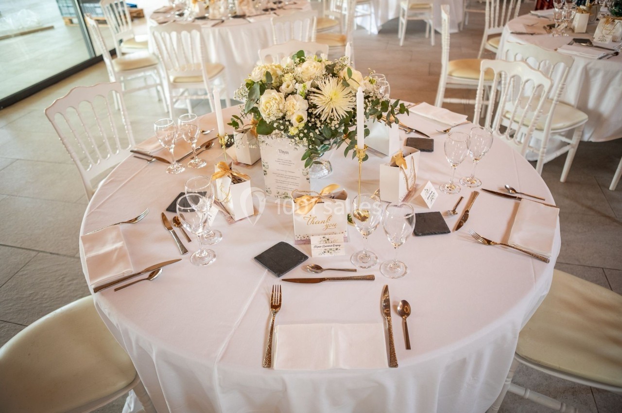 Table ronde élégamment dressée avec nappes blanches, couverts dorés, centre de table floral et cadeaux invités.