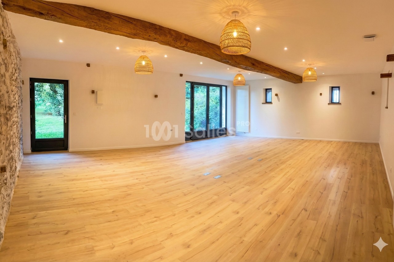 Salle lumineuse avec parquet en bois clair, murs blancs, poutres apparentes et grandes fenêtres donnant sur un jardin.