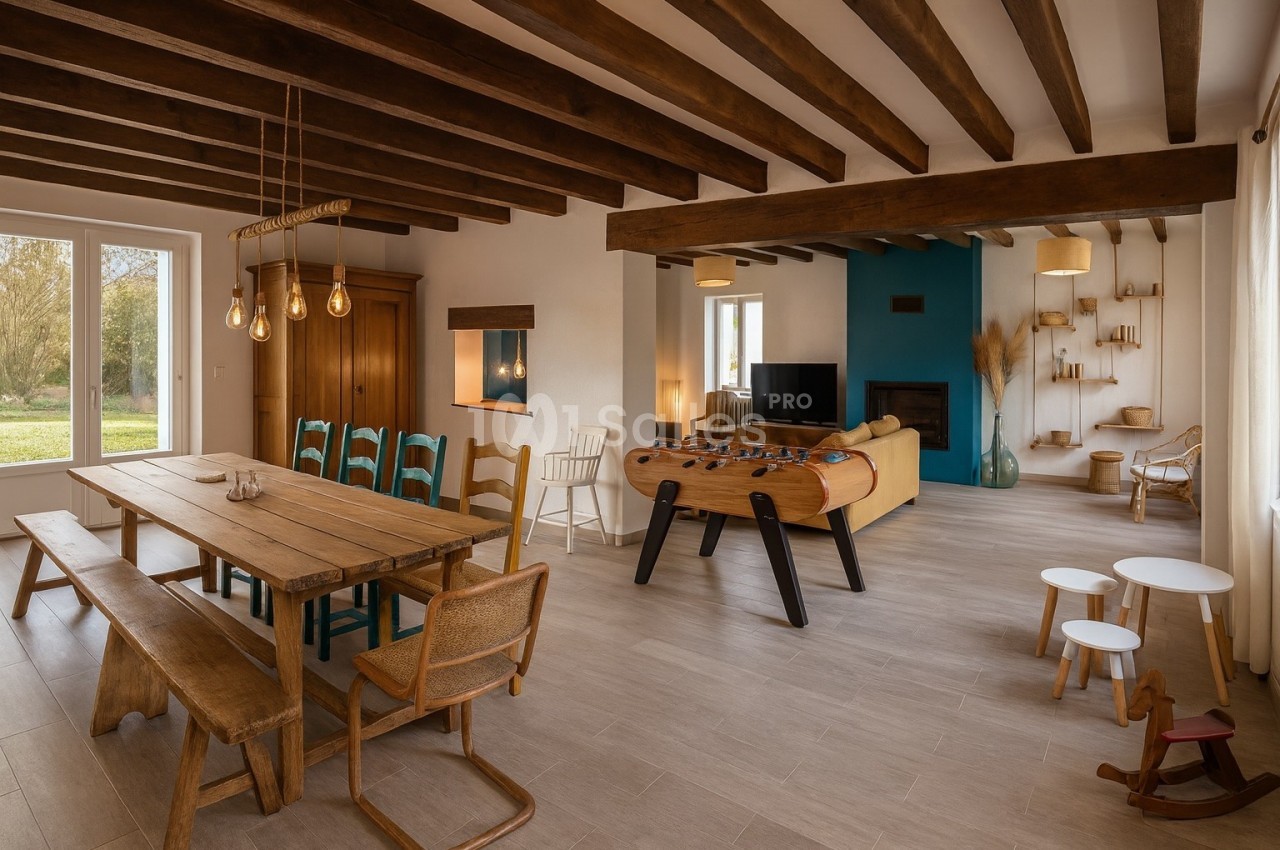 Salle de séjour lumineuse avec table en bois, baby-foot, canapé jaune, poutres apparentes et vue sur un jardin.