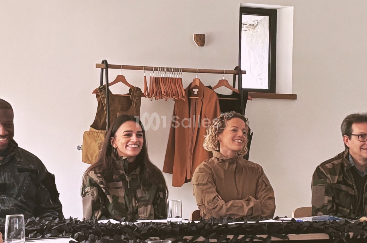 Des personnes en tenue militaire assises autour d'une table, souriantes, avec des vêtements suspendus en arrière-plan.