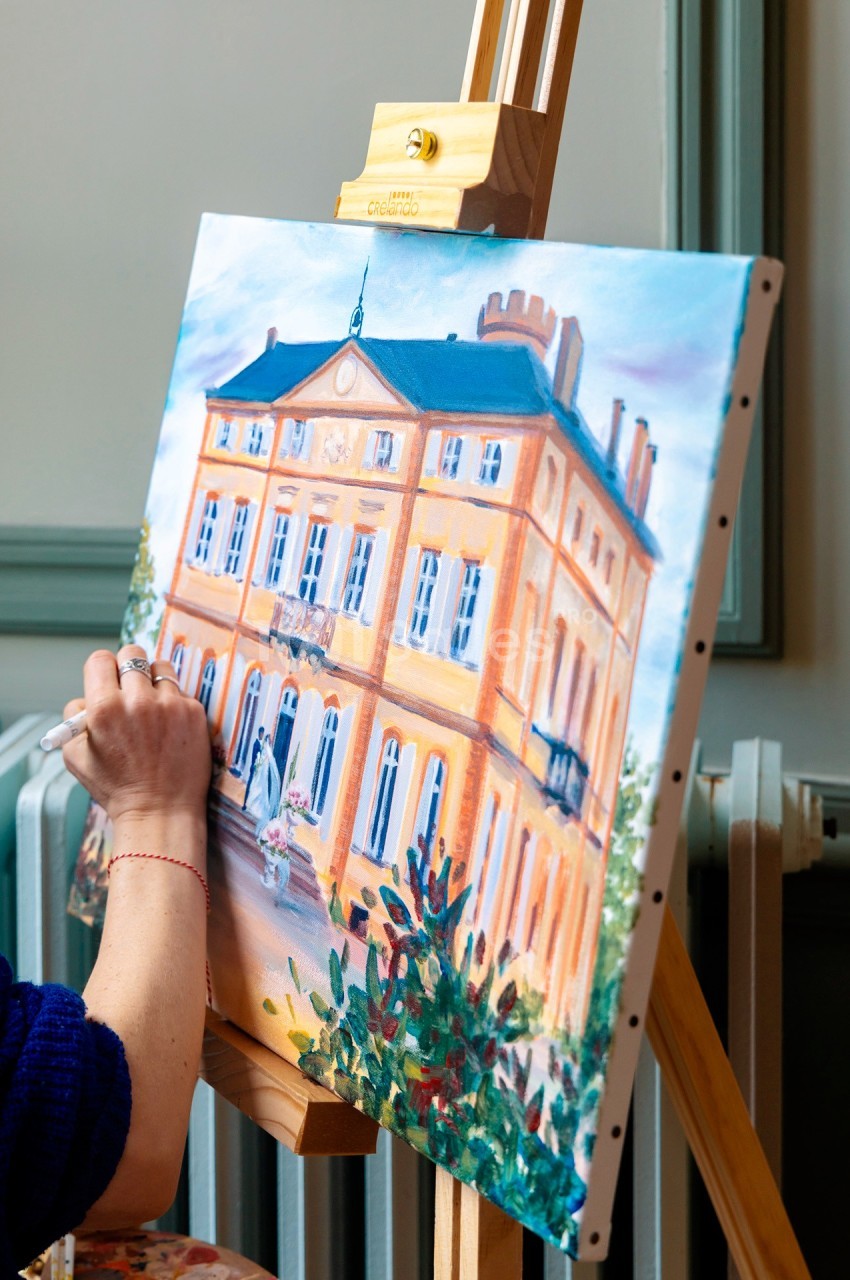 Personne peignant un tableau représentant un bâtiment jaune avec des détails architecturaux, posé sur un chevalet.