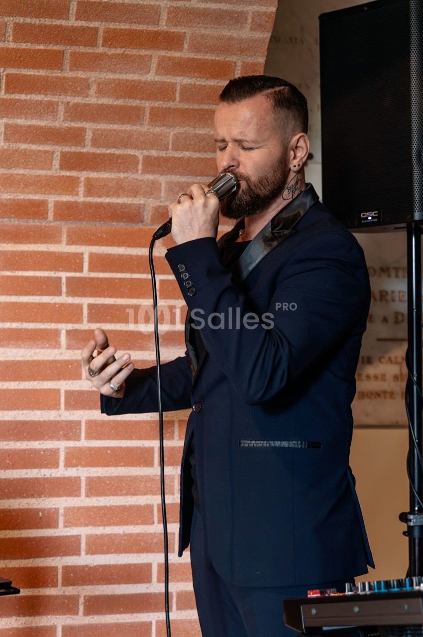 Un homme en costume sombre chante au micro devant un mur en briques, avec un haut-parleur en arrière-plan.