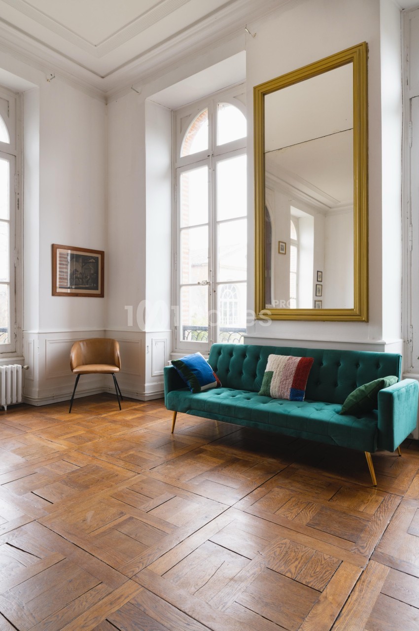 Salon lumineux avec parquet en bois, canapé vert, fauteuil en cuir, grand miroir doré et fenêtres hautes.