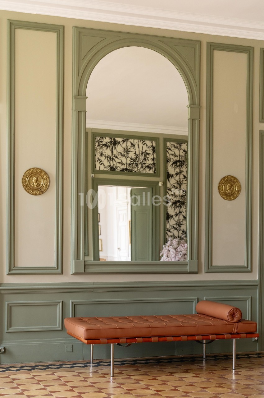 Banc en cuir brun devant un grand miroir encadré de moulures vertes, dans une pièce au sol en carreaux anciens.