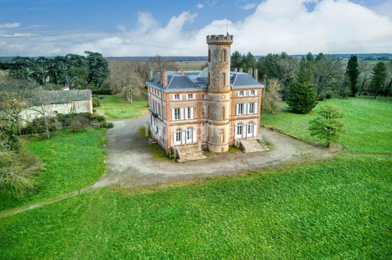Château en briques rouges avec une tour centrale, entouré de pelouses et d'arbres, sous un ciel partiellement nuageux.