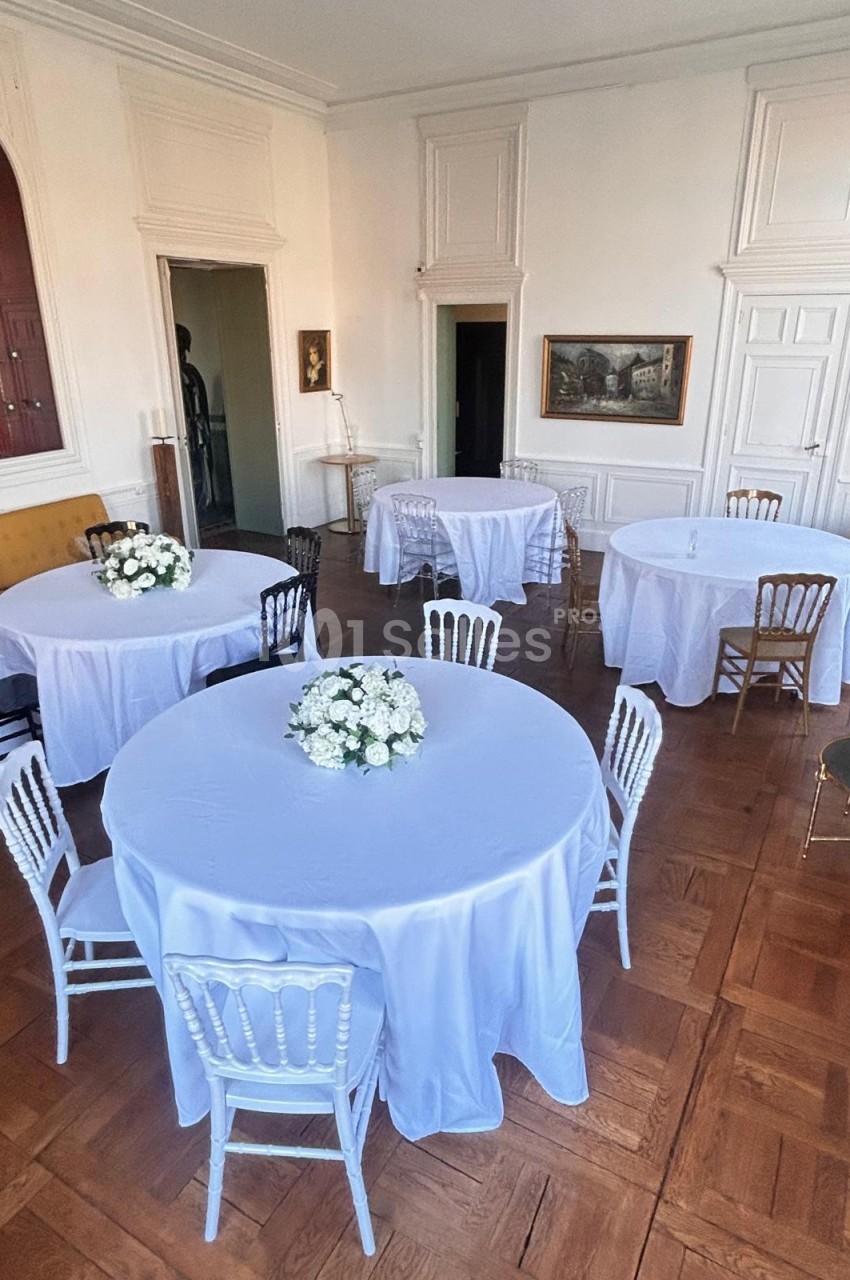 Salle lumineuse avec tables rondes recouvertes de nappes blanches, décorées de bouquets de fleurs blanches.