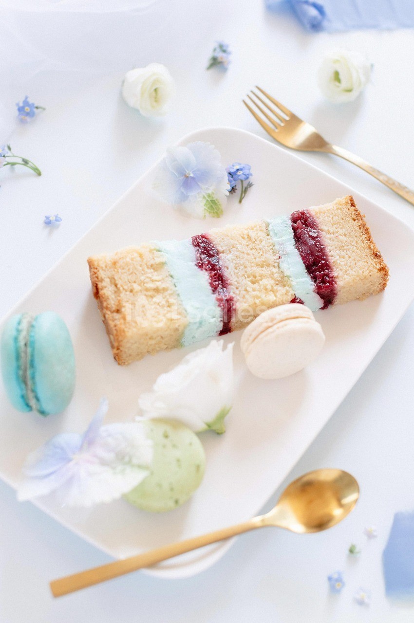 Assiette avec une tranche de gâteau décorée de crème bleue, macarons colorés et fleurs en fond.