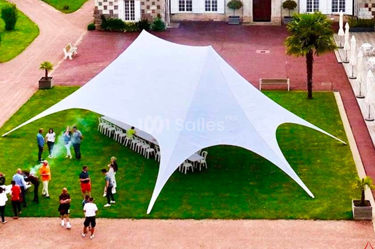Tente blanche montée sur une pelouse, entourée de personnes et de tables, dans une cour avec bâtiments en arrière-plan.