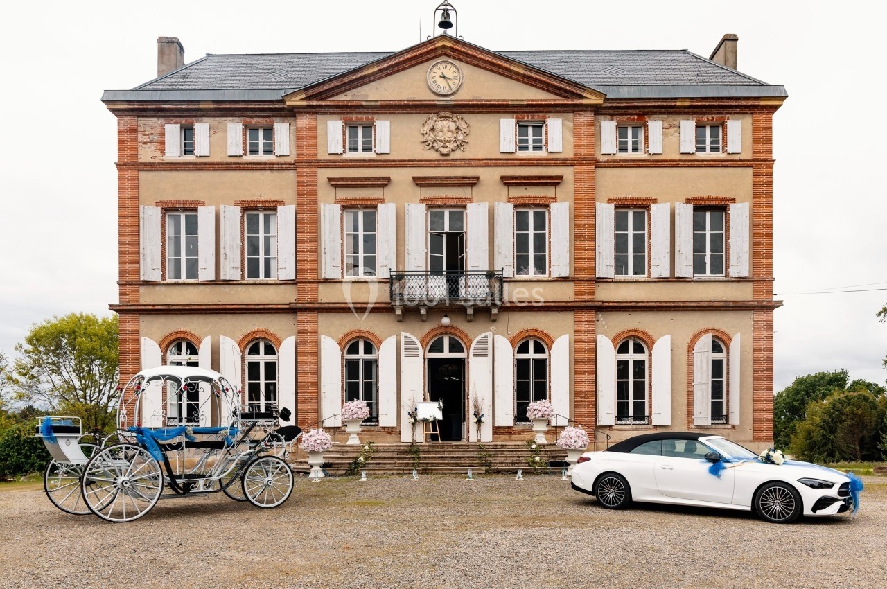 Façade d'un château en briques avec une calèche et une voiture blanche stationnées devant.