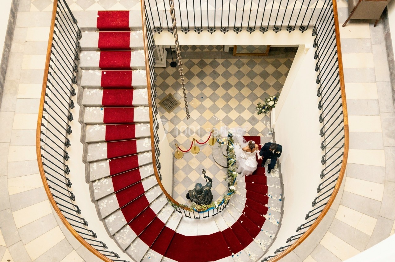 Vue plongeante d'un escalier en colimaçon avec tapis rouge, un couple en tenue de mariage au rez-de-chaussée.