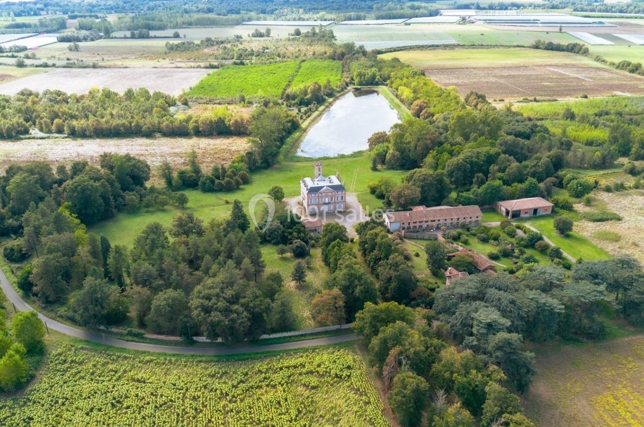 Vue aérienne d'un domaine avec un château, des bâtiments annexes, un étang et des champs environnants.