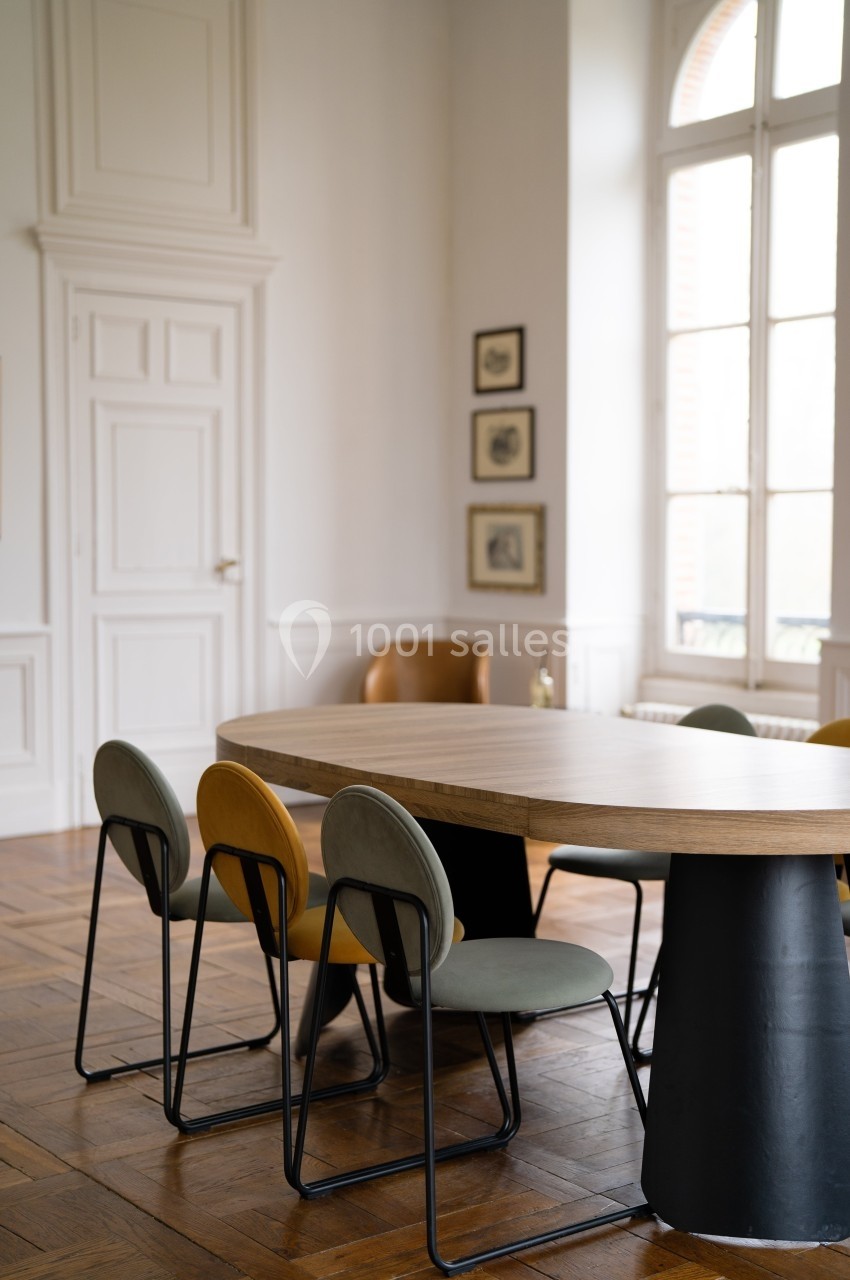 Table en bois entourée de chaises modernes dans une pièce lumineuse avec parquet et grandes fenêtres.