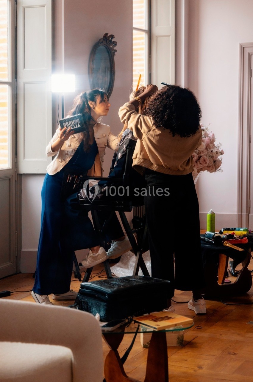 Deux personnes maquillent une femme assise sur une chaise dans une pièce lumineuse avec des fenêtres ouvertes.