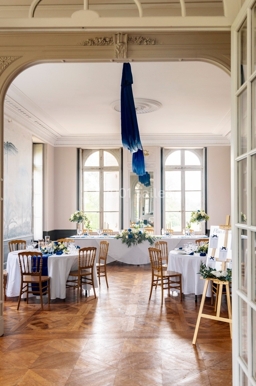 Salle élégante avec tables rondes dressées, nappes blanches, décorations florales et grandes fenêtres lumineuses.