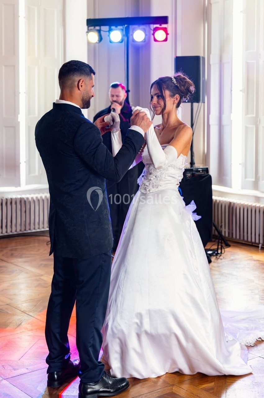 Un couple en tenue de mariage danse dans une salle éclairée, accompagné d’un musicien jouant du violon.