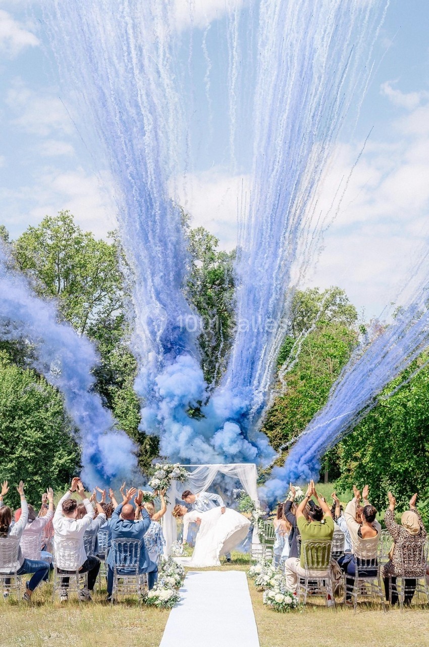 Des invités applaudissent lors d'une cérémonie en plein air avec des fumées bleues s'élevant dans le ciel.