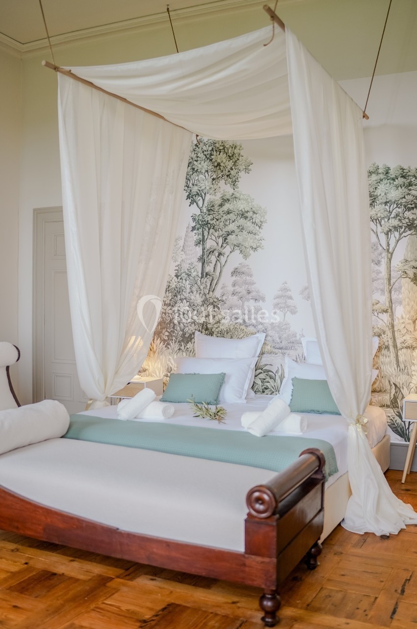 Lit à baldaquin avec drapés blancs, literie pastel et décor mural représentant une forêt, dans une chambre lumineuse.