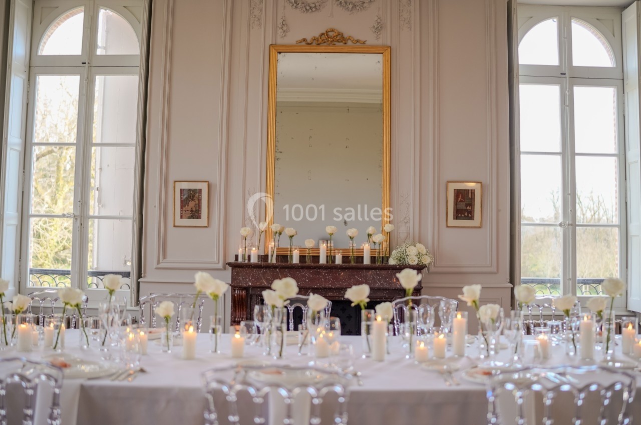 Table élégamment dressée avec des bougies et des fleurs blanches dans une salle lumineuse aux grandes fenêtres et miroir…