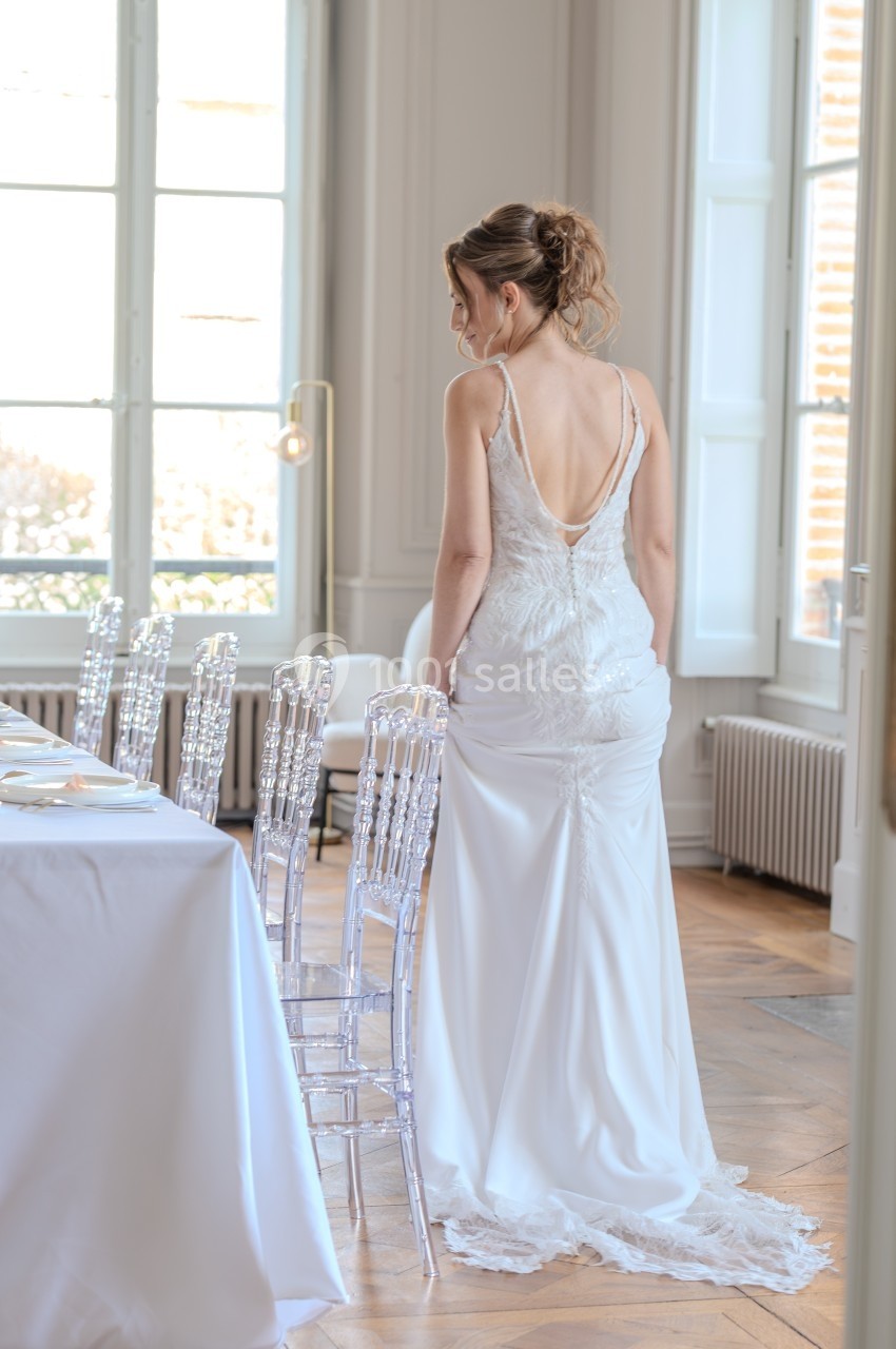 Une femme en robe de mariée blanche, de dos, se tient près d'une table dressée dans une pièce lumineuse.