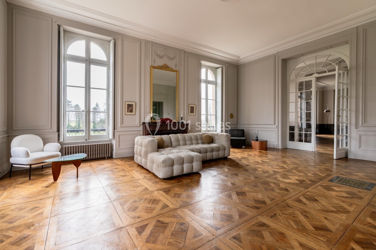 Salon lumineux avec parquet en bois, canapé beige, fauteuil, table basse et grandes fenêtres donnant sur un jardin.
