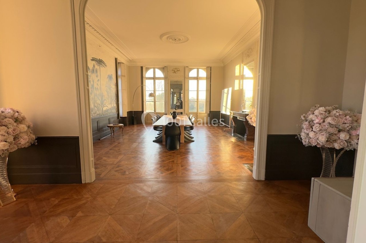 Salle lumineuse avec parquet en bois, grande table noire centrale, murs ornés et bouquets de fleurs roses.