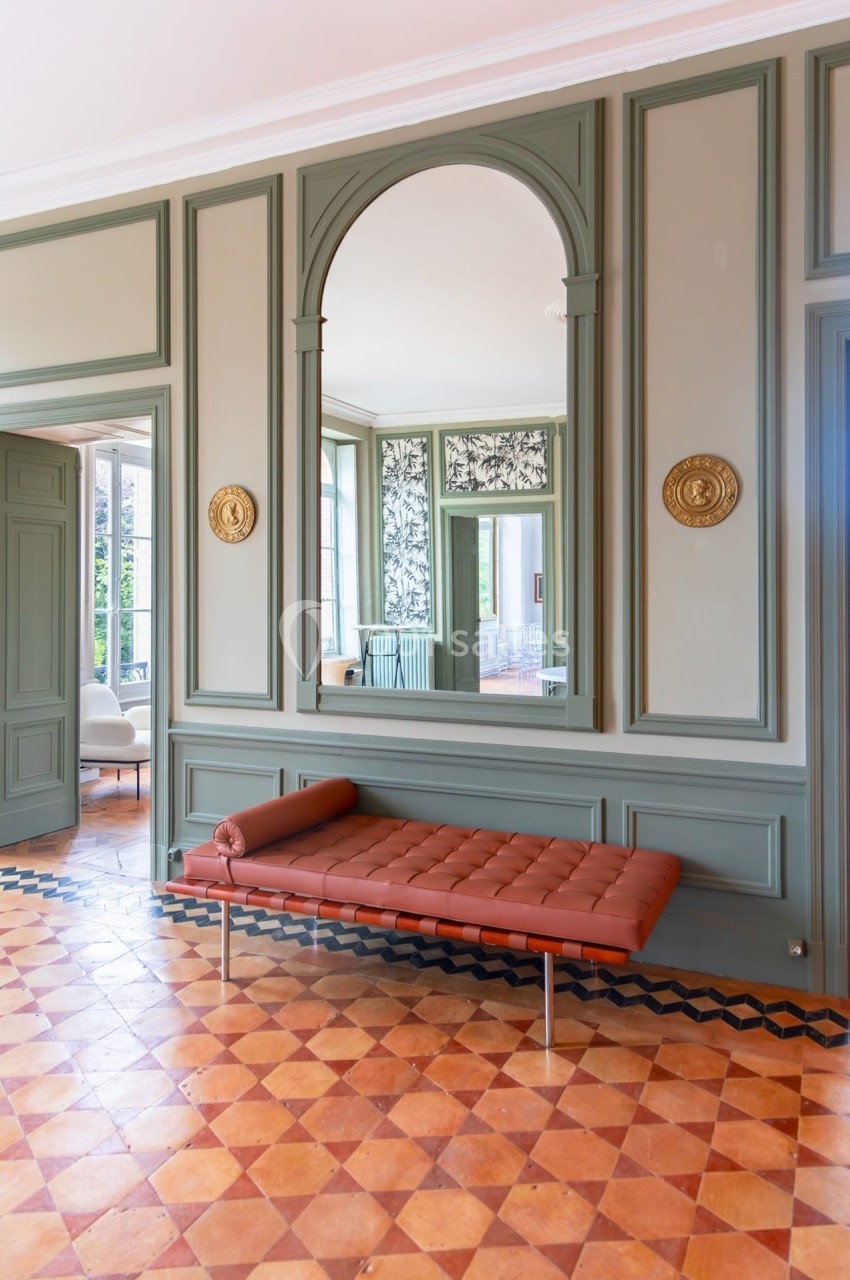 Banc en cuir marron devant un grand miroir encadré de moulures vertes dans une pièce lumineuse au sol en carreaux.