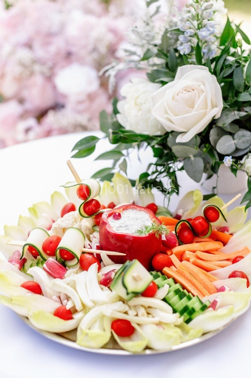 Plateau de crudités variées avec une sauce au centre, décoré de fleurs blanches en arrière-plan.