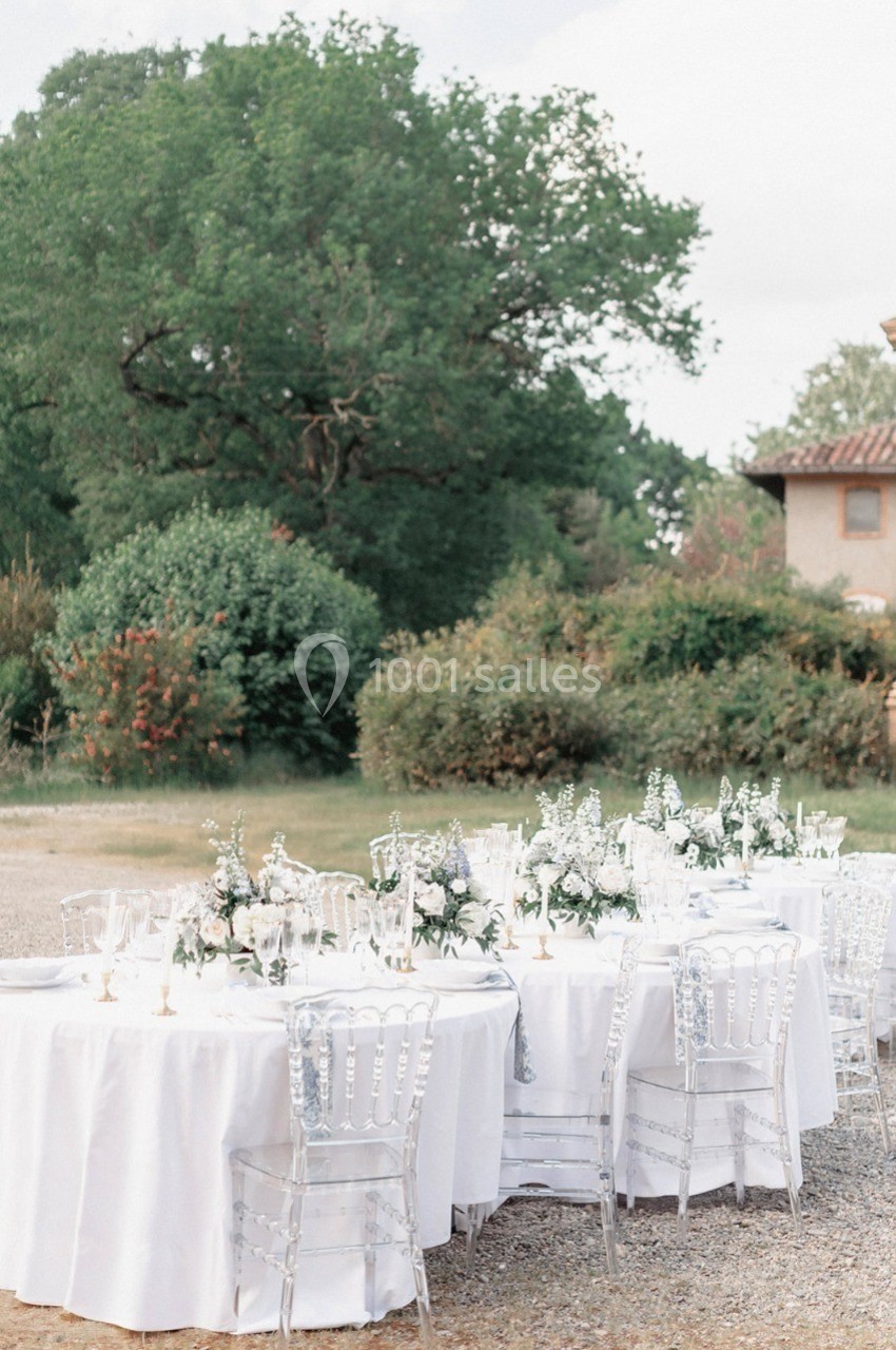 Tables rondes dressées avec nappes blanches, décorées de fleurs, disposées en extérieur près d'arbres et d'un bâtiment.