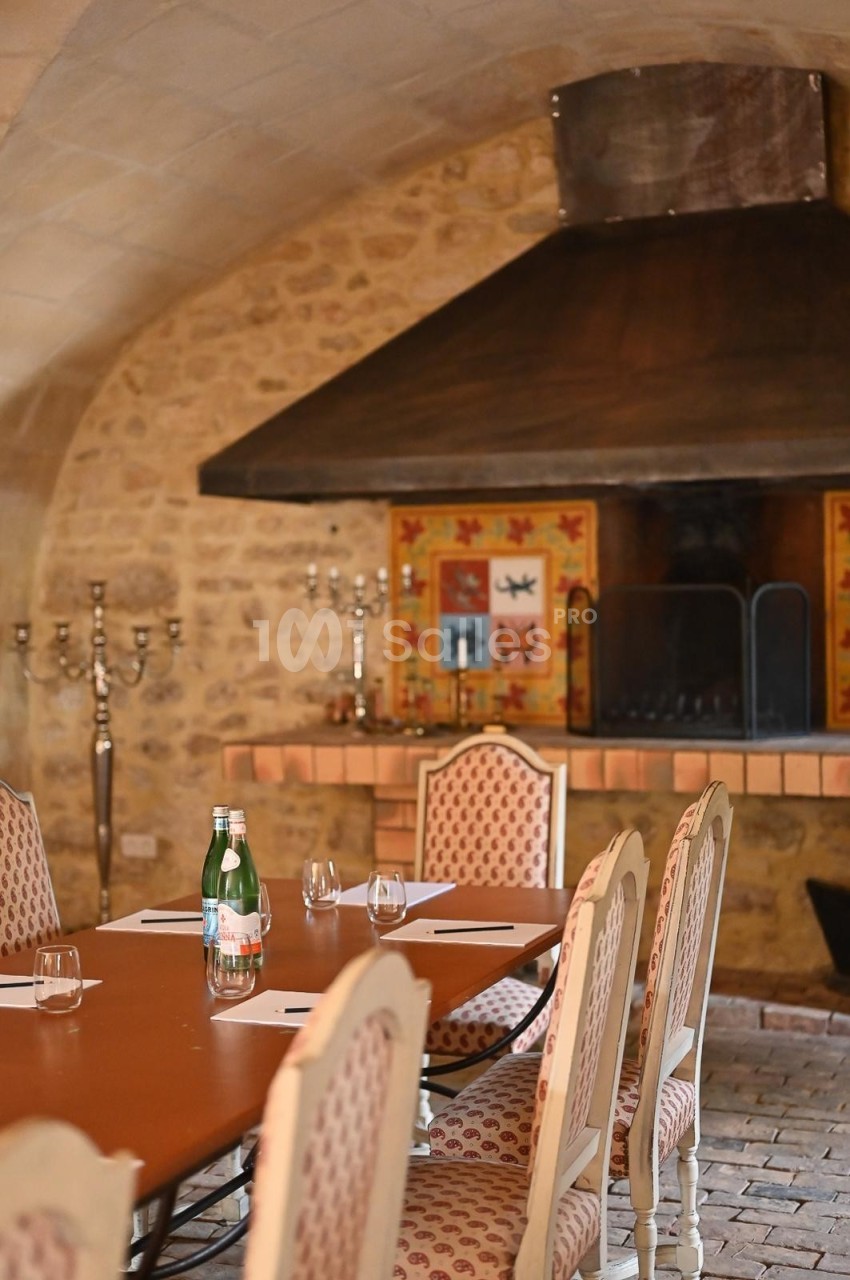 Salle en pierre avec une grande cheminée, table ronde dressée avec chaises rembourrées et bouteille d'eau.