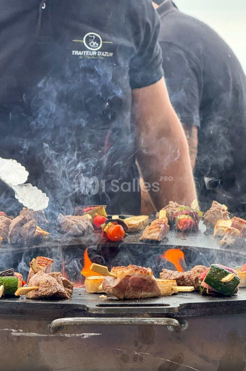Viande et légumes grillés sur une plancha en extérieur, avec un cuisinier en arrière-plan.