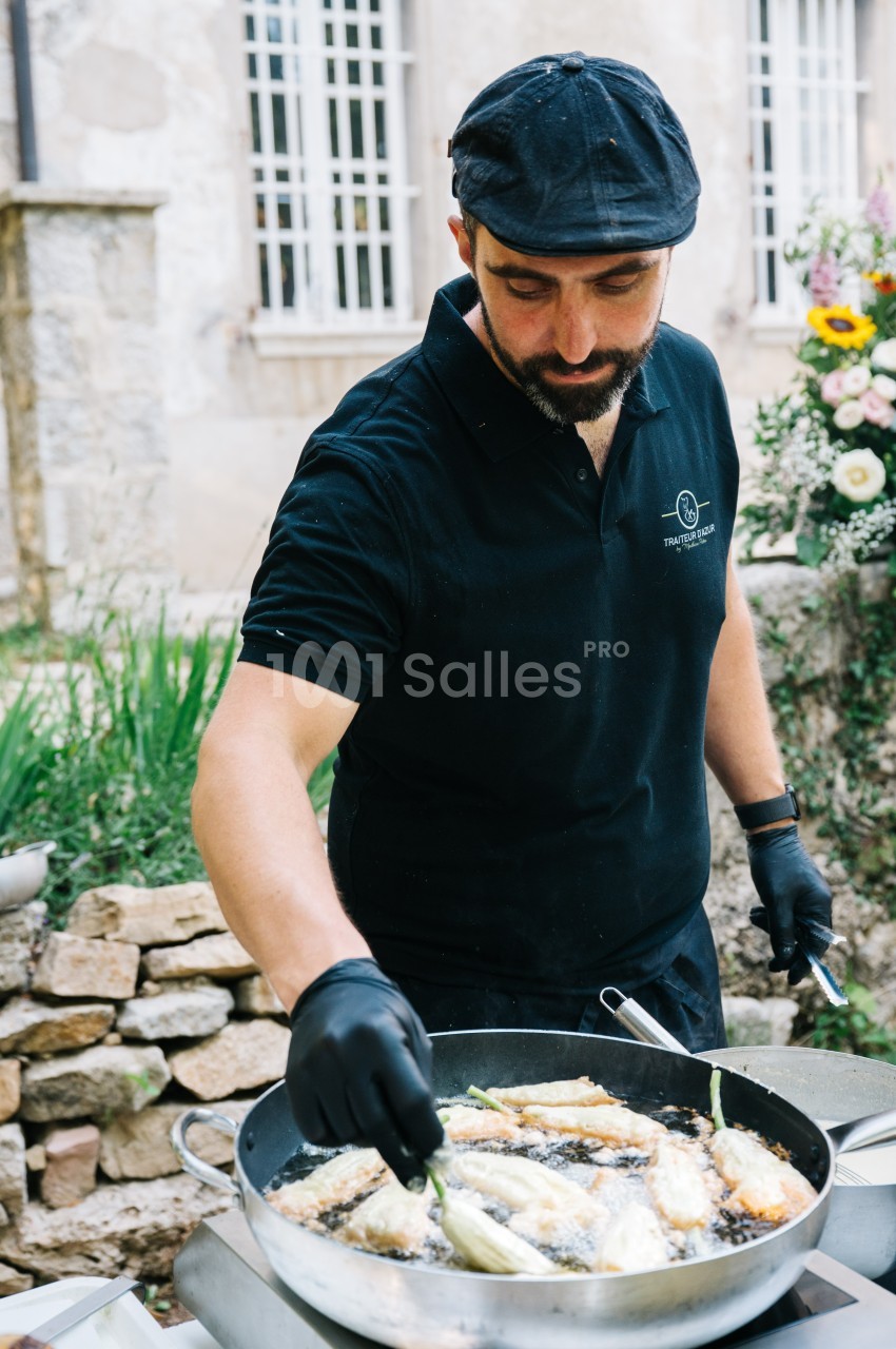 Un cuisinier en tenue noire prépare des légumes dans une grande poêle en extérieur, devant un mur en pierre.