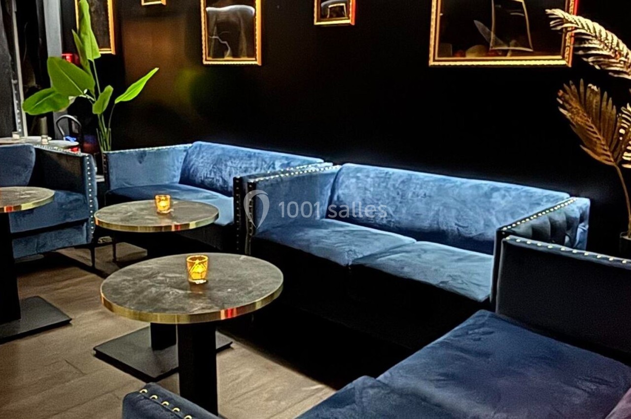 Canapés bleus en velours entourant des tables rondes noires avec bougies, dans un intérieur sombre et élégant.