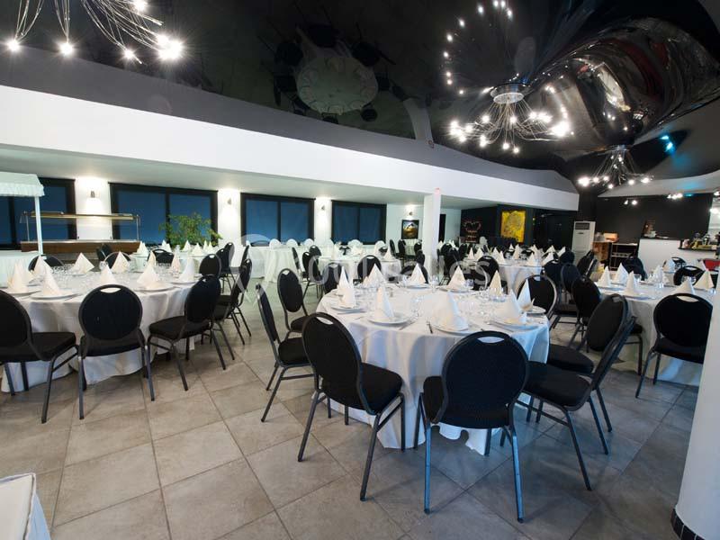 Salle de réception avec tables rondes dressées, nappes blanches et chaises noires, éclairage moderne au plafond.