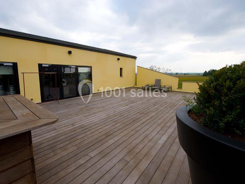 Terrasse en bois avec un bâtiment jaune, des transats et une grande jardinière sous un ciel nuageux.