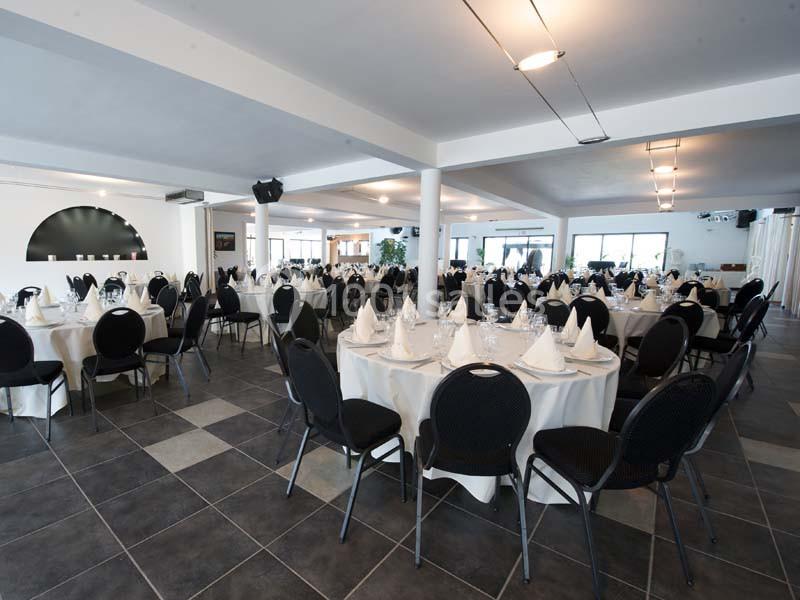 Salle de réception avec tables rondes dressées, nappes blanches, chaises noires et éclairage naturel.
