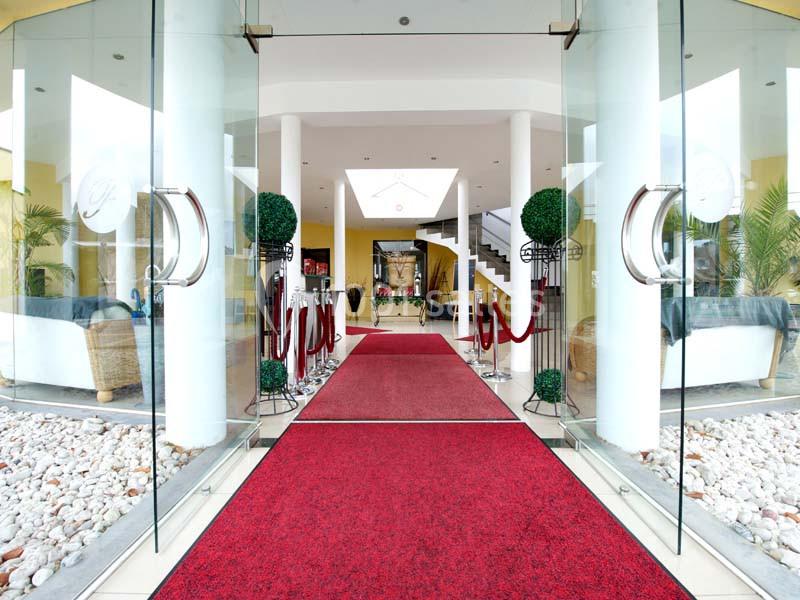 Entrée d'un bâtiment avec tapis rouge, portes vitrées ouvertes, colonnes blanches et décoration végétale.