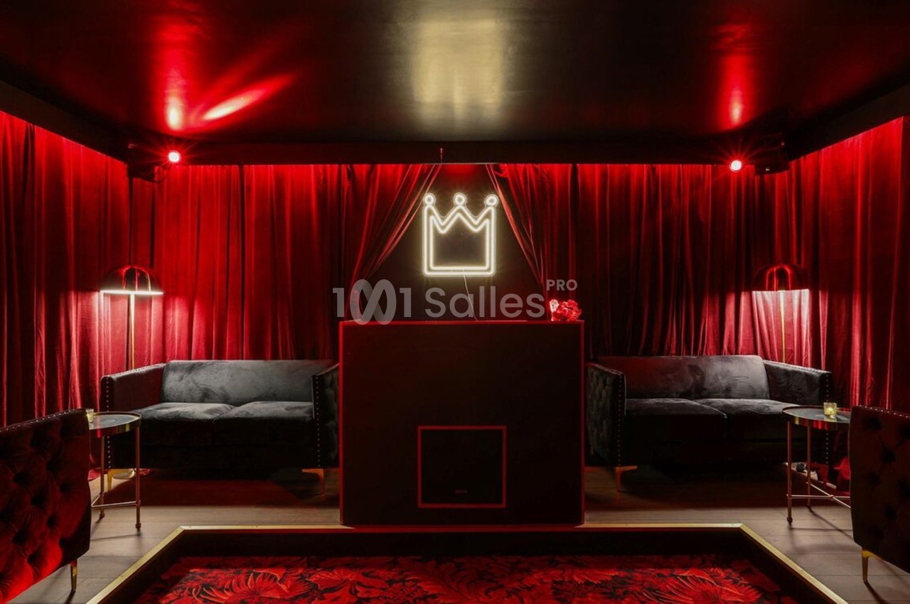 Salle sombre avec rideaux rouges, canapé noir, éclairage tamisé et enseigne lumineuse en forme de couronne.