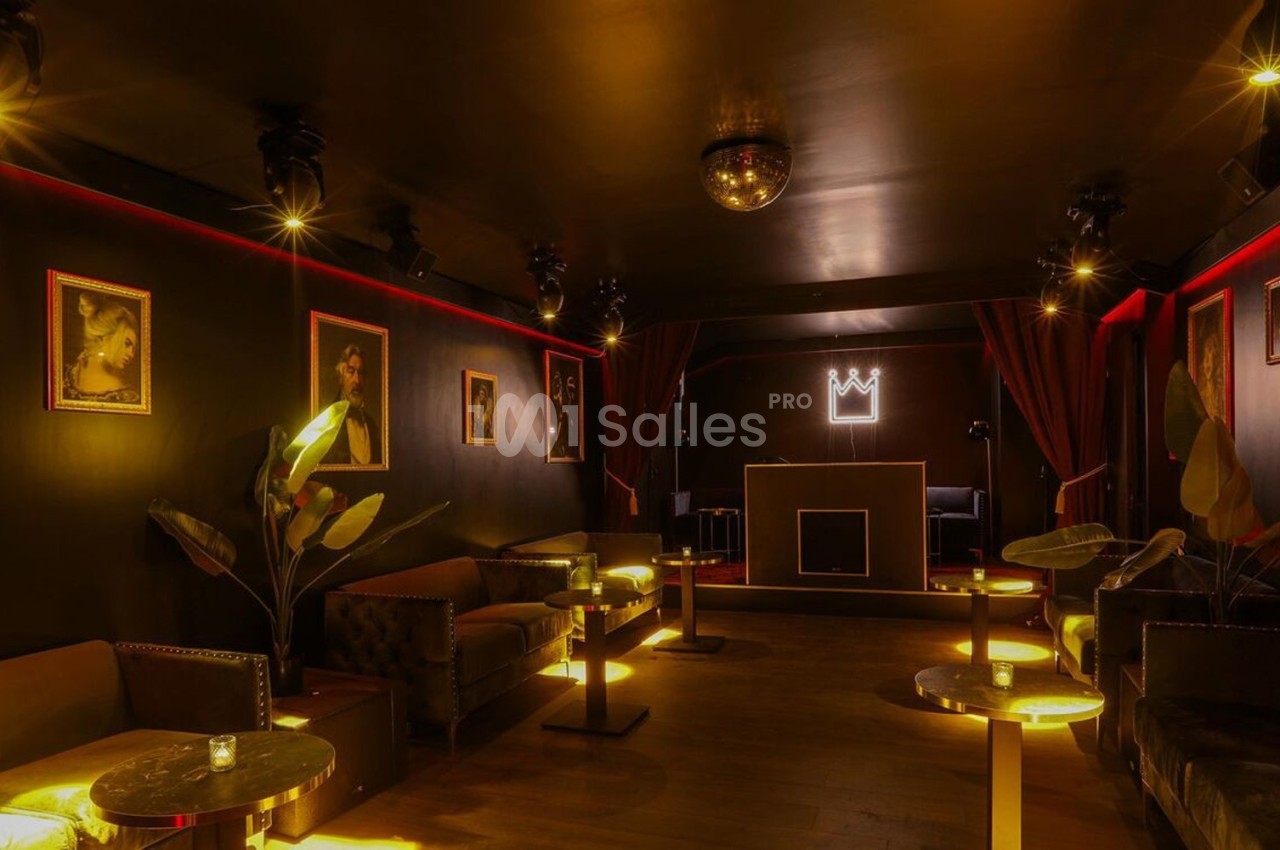Salle sombre et élégante avec canapés, tables éclairées, portraits aux murs et une cabine de DJ au fond.