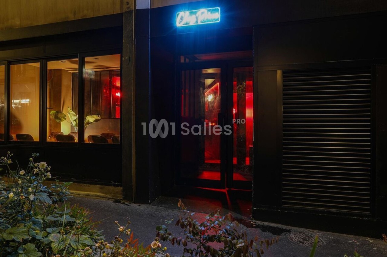 Façade d'un établissement avec une entrée vitrée, éclairée par une lumière rouge et une enseigne lumineuse bleue.