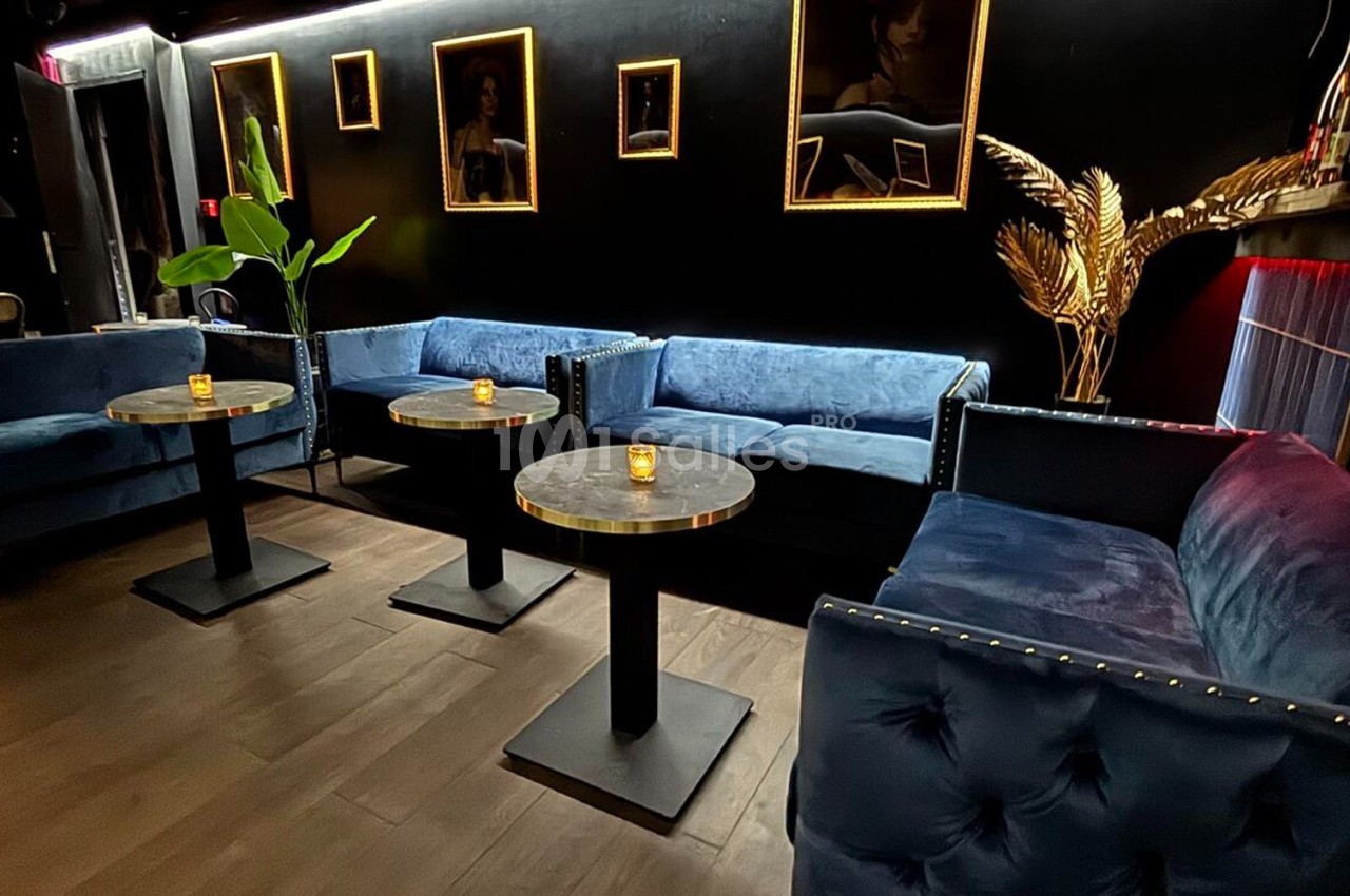 Canapés bleus en velours, tables rondes avec bougies, murs noirs décorés de cadres dorés et éclairage tamisé.