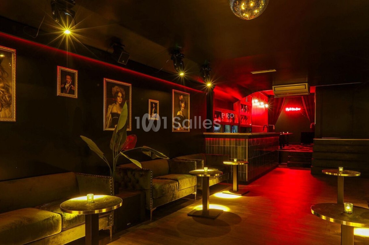 Salle de bar intimiste avec éclairage tamisé rouge, canapés, tables basses et portraits encadrés sur les murs.