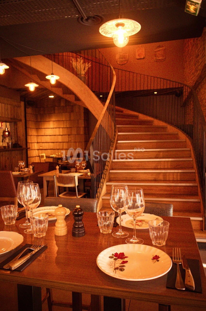 Table dressée avec vaisselle et verres dans un restaurant chaleureux, avec escalier en bois en arrière-plan.