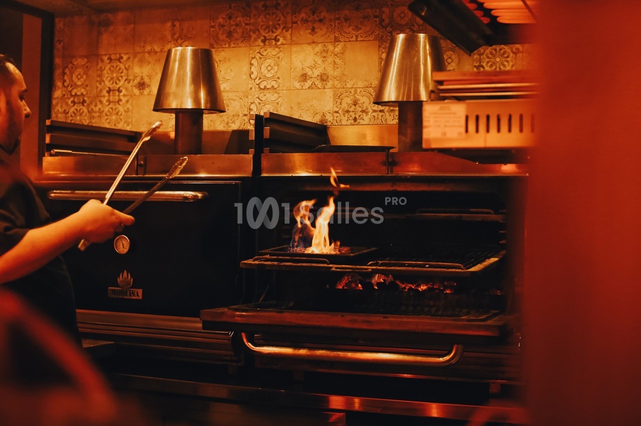 Un cuisinier manipule des pinces devant un four ouvert avec des flammes visibles sur un gril.