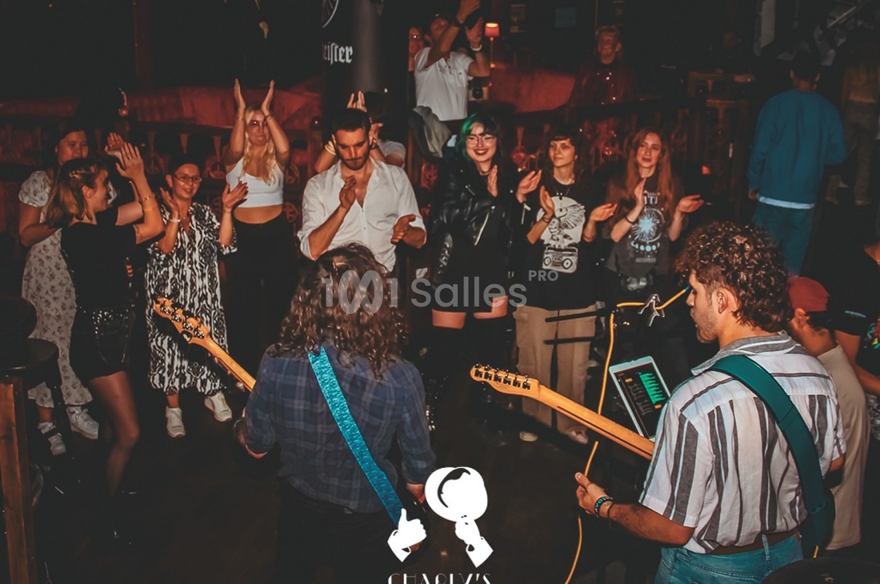 Musiciens jouant sur scène dans un bar, entourés d'un public debout applaudissant et dansant.